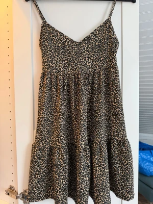 Leopardmönstrad kort klänning - Säljer en snygg kort klänning med leopardmönster och tunna axelband. Klänningen har en v-ringad urringning och är tillverkad i ett lätt material som passar perfekt för varma dagar. Den har en avslappnad passform och volang nedtill.