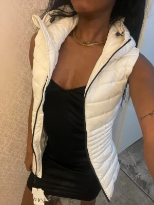 Vit dunväst med hög krage - Snygg vit dunväst från Only med hög krage och dragkedja framtill. Västen har en figurnära passform och quiltad design. OBS! Helt ny med prislapp. Västen har en fläck (smink - rouge) på vänster sida som förmodligen går bort i en ordentlig tvätt. Pga fläcken är priset ordentligt refuserat!