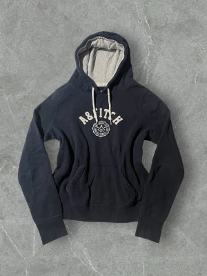 Mörkblå hoodie från Abercrombie & Fitch | 40 - Mörkblå hoodie med vit broderad logga och text på bröstet från Abercrombie & Fitch. Utrustad med huva, vita dragsnören och känguruficka. Insidan av huvan är ljusgrå för en snygg kontrast. Perfekt för en avslappnad och cool stil.
