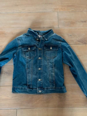 Blå jeansjacka från Sincerely Loved - Klassisk blå jeansjacka från Sincerely Loved med raka sömmar, två bröstfickor med knappar och snygga metallknappar framtill. Jackan har en tidlös passform och är perfekt att slänga över en hoodie eller t-shirt för en chill look.