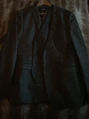 Mörkgrå klassisk kostym  - Snygg mörkgrå kavaj med två framfickor och bröstficka. Enkelknäppt modell med smal krage och diskret struktur i tyget. Passar perfekt till skjorta eller t-shirt för en stilren look. Materialet ser ut att vara en blandning av ull och syntet.
