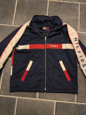 Tommy Hilfiger Vintage Jacka från 90 talet - Snygg retro vindjacka/ vår jacka från Tommy Hilfiger i marinblått, vitt och rött. Jackan har dragkedja framtill, stora loggor på både fram- och baksida samt coola färgblock och detaljer längs ärmarna. Perfekt för dig som gillar streetwear och klassisk 90-talsstil. Helt fri från märken eller skador.  Väldigt sällsynt med en sån gammal jacka utan några defekter.