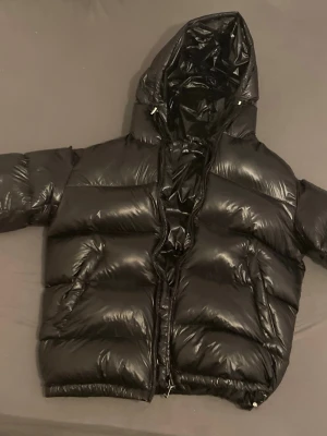 Zara puffer jacka - Säljer en svart, glansig pufferjacka från Zara  med stor huva och dragkedja framtill. Jackan har breda, vadderade paneler och elastisk nederkant för extra komfort. Perfekt för kalla vinterdagar och riktigt snygg streetstil.