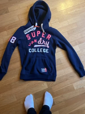 Marinblå Superdry hoodie college - Snygg marinblå hoodie från Superdry med stora tryck i vitt, rosa och rött på bröstet och siffran 68 på ärmen. Klassisk huva med vita snören och känguruficka framtill. Perfekt för en avslappnad och sportig stil. Mjuk bomullsblandning. Denna e en av de skönaste tröjor jag har haft från superdy jag e galet imponerad men som e synd e att den e för liten.Skulle säga att denna passar till nån som e ca 178 den e lite liten på armarna och lämgden och jag e 182 . 