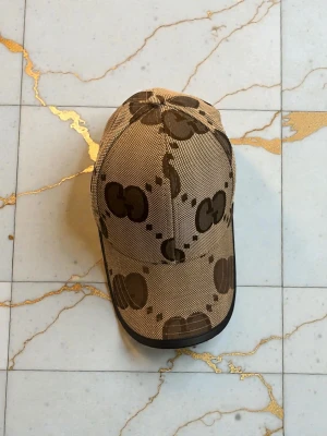 Beige keps med GG-mönster från Gucci - Säljer en beige keps från Gucci med det klassiska GG-mönstret i brunt. Kepsen har böjd skärm och är tillverkad i ett slitstarkt tygmaterial. Perfekt för dig som vill ha en stilren accessoar med lyxig känsla och tydlig logga.