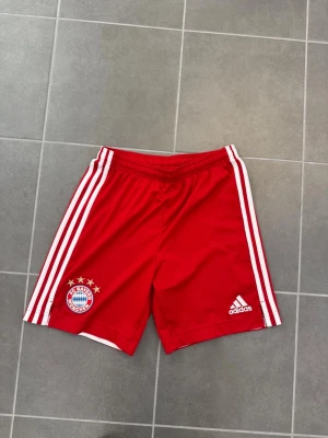 Bayern München shorts Adidas röda - Röda Bayern München shorts från Adidas med vita ränder längs sidorna och klubbens logga på ena benet. Perfekta för fotbollsträning eller match. Tillverkade i lätt och svalt material för bästa komfort.