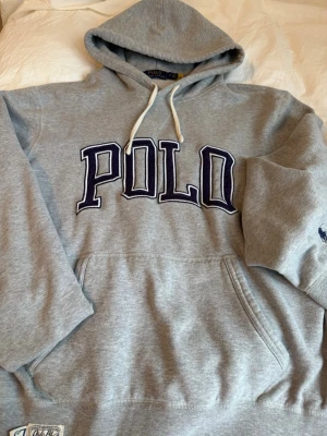 Grå vintage hoodie från Polo Ralph Lauren - Snygg grå hoodie från Polo Ralph Lauren med stor POLO-text i mörkblått framtill och klassiska loggan broderad på ärmen. Hoodien har huva med vita snören och en stor magficka. Tillverkad i mjuk bomullsblandning, perfekt för en chill och avslappnad stil.