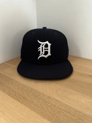 Svart New Era keps 54,9 cm (aldrig använd) - Säljer en svart keps från New Era med rak skärm och broderad vit D-logga framtill. Klassisk snapback-modell med MLB-logga bak och New Era-logga på sidan. Tillverkad i bomullsmix, perfekt för en streetwear-look.
