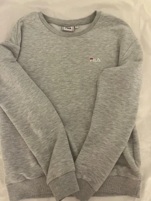 Grå sweatshirt från FILA 💞 - Klassisk grå sweatshirt från FILA med rund halsringning och diskret logga broderad på bröstet. Tröjan har långa ärmar och ribbade muddar vid ärmslut och nederkant. Perfekt för en avslappnad och sportig stil. Det finns ett litet hål på tröjan men inget man direkt märker💞