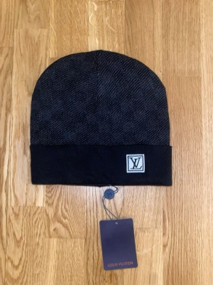 Svart Mössa - Snygg svart mössa med klassiskt rutmönster och vikt kant. Mössan har en diskret logga framtill och är tillverkad i mjukt, stickat material som känns lyxigt och bekvämt.