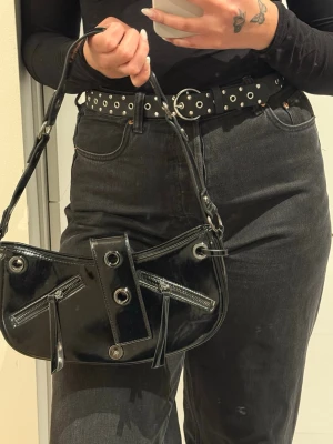 Svart lack handväska med nitar - Snygg svart handväska i glansigt lackmaterial med silverfärgade metalldetaljer och nitar. Väskan har två dragkedjor framtill, dekorativa öljetter och ett brett axelband. Perfekt för dig som gillar en edgy och trendig stil. Det fattas magneten för att stänga väskan!