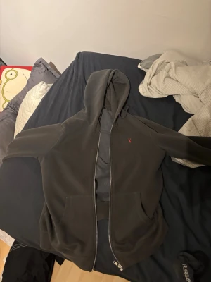 Mörkgrå hoodie med dragkedja - Säljer en mörkgrå hoodie med dragkedja och huva. Den har en liten röd broderad logga med hjorthuvud på bröstet och två stora fickor framtill. Perfekt för chill dagar och enkel att matcha med andra plagg.