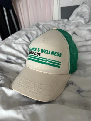 Grön och beige keps - Snygg keps i beige och grönt med texten 'Balance & Wellness Athletic Club' framtill. Kepsen har klassisk böjd skärm och mesh-tyg baktill för extra ventilation. Perfekt för en sportig och avslappnad stil.