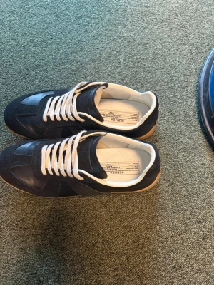 Mason Margiela skor - Snygga svarta sneakers från Mason Margiela med vita snören och beige innersula. Skorna har en klassisk låg siluett, ovandel i mocka och läder samt en brun gummisula. Perfekta för dig som gillar stilrena och tidlösa sneakers.