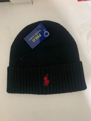 Svart mössa från Polo Ralph Lauren - Snygg svart ribbstickad mössa från Polo Ralph Lauren med klassisk röd logga broderad framtill. Tillverkad i mjukt material som håller dig varm och stilren under kalla dagar. Perfekt accessoar för dig som vill ha en enkel men ikonisk look.