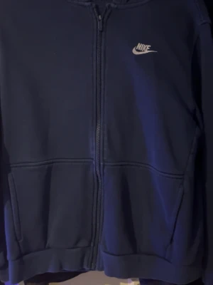 Mörkblå zip hoodie från Nike - Snygg mörkblå hoodie från Nike med dragkedja framtill och klassisk vit logga på bröstet. Hoodien har två fickor och en skön huva. Perfekt för chill dagar eller när du vill ha en sportig look. Tillverkad i mjukt material för extra komfort.