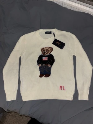 Vit stickad tröja Polo Bear RL - Vit stickad tröja från Polo Ralph Lauren med ikoniska Polo Bear-motivet framtill. Björnen har på sig jeans och svart tröja med amerikanska flaggan. RL-broderi i rött nedtill. Rund halsringning och ribbade muddar. Perfekt statement-plagg för dig som gillar klassisk stil.