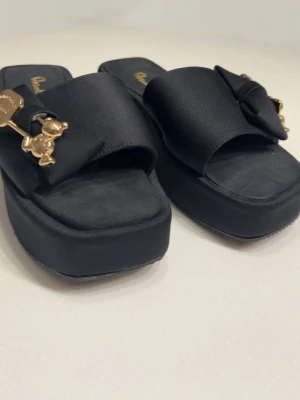 Svarta platåsandaler med rosett - Stilrena svarta sandaler från bimbo med breda band i satinliknande material och stor rosett framtill. Dekorerade med guldfärgade berlocker och logga. Platåsula och fyrkantig tå ger en trendig look. Perfekta för dig som gillar statement-detaljer.