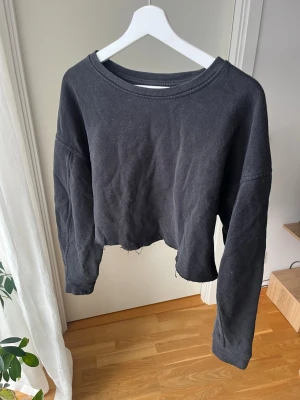 Svart croppad sweatshirt - En svart croppad sweatshirt med rå kant nertill och långa ärmar. Tröjan har en avslappnad passform och rund halsringning. Perfekt för en chill och trendig look. 
