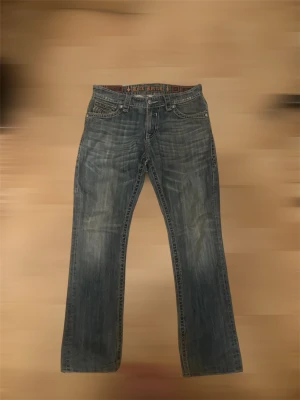 rock revival bootcut jeans 32 john  - Säljer ett par blå bootcut jeans från Rock Revival med slitningar och broderade detaljer på bakfickorna. Jeansen har klassisk femficksmodell, snygga kontrastsömmar och är tillverkade i denim. Perfekta för dig som gillar en avslappnad men ändå trendig stil.