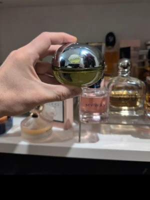 DKNY Be Delicious parfym äppelflaska - Fräsch parfym från DKNY i den ikoniska Be Delicious-flaskan som liknar ett grönt äpple. Flaskan är rund med en blank silverfärgad topp och grön glasbotten. Doften är känd för sina fruktiga och fräscha toner, perfekt för dig som gillar en energisk vibe.