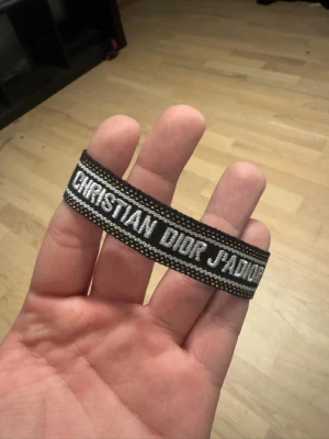 Christian Dior J'adior tygarmband - Snyggt svart tygarmband från Christian Dior med texten 'J'adior' broderad i vitt. Armbandet har dekorativa detaljer i grått och vitt samt tofsar i ändarna. Perfekt accessoar för dig som gillar exklusiva märken och vill sticka ut.