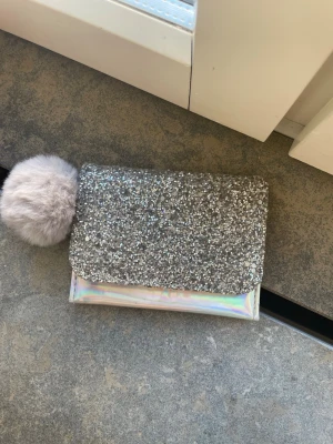 Glittrig silver kuvertplånbok med fluffboll - Säljer en superfin plånbok med glittrigt silverlock och holografisk kropp. Väskan har en stor, mjuk grå fluffboll som detalj. Perfekt för dig som vill sticka ut och addera lite extra bling till din outfit. Ingen synlig logga eller märke.