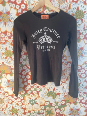 Brun långärmad topp Juicy Couture - Brun långärmad topp från Juicy Couture med vit text och krona framtill. Modellen är figurnära och har rund halsringning. Materialet känns mjukt och stretchigt, troligen bomull eller bomullsblandning. Perfekt för dig som gillar en klassisk men ändå statement look.