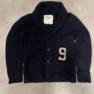 Mörkblå cardigan från Abercrombie & Fitch - Mörkblå stickad kofta från Abercrombie & Fitch med bred och stora knappar framtill. Snygg detalj med en vit broderad siffra 9 på bröstet. Ribbstickade muddar vid ärmslut och krage ger en extra stilren look.