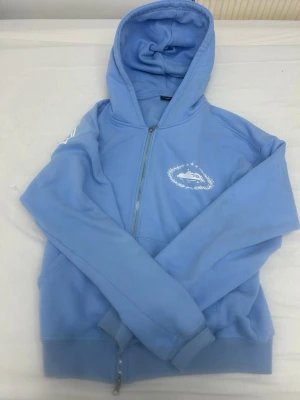 Blå zip hoodie från Corteiz - Snygg ljusblå hoodie från Corteiz med dragkedja framtill och stor vit logga på ryggen. Har huva och ribbade muddar vid ärmar och midja. Liten vit logga på bröstet och detaljer i vitt. Perfekt för en avslappnad streetwear-look.