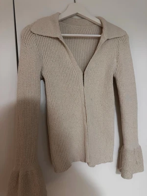 Aprikos ribbstickad kofta  - Trendig aprikos ribbstickad kofta med bred krage och djup v-ringning. Koftan har långa ärmar med snygga volangdetaljer vid ärmsluten. Tillverkad i 100% polyester för en mjuk och stretchig känsla. Perfekt för dig som gillar en clean och stilren look.