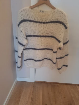 Vit stickad tröja med blå ränder - Mysig vit stickad tröja med breda mörkblå horisontella ränder. Tröjan har en avslappnad passform, rund halsringning och långa ärmar. Perfekt för dig som gillar en enkel men snygg stil. Materialet är tjockt och fluffigt för extra komfort.