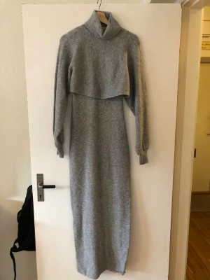 Grå stickad långklänning polo - 2-delad. Mysig grå stickad långklänning med andra delen som är avtagbar, cool tröjdel med polokrage och långa ärmar. Klänningen har en relaxed passform och ribbade muddar vid ärmslut och hals. Perfekt för kyligare dagar och enkel att styla med boots eller sneakers.
