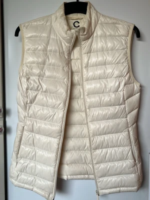 Krämvit dunväst  - Snygg krämvit dunväst från C i modellen Super Light Down. Tillverkad i lätt och smidigt material och varm med äkta dun i. Det håller värmen bra för vid alla tillfällen. Skulle ändå säga storlek S Funkar för ute promenader, träning eller annan aktivitet 🌟🌸