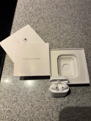 Apple AirPods med laddningsetui - Säljer ett par vita Apple AirPods med tillhörande laddningsetui och originalförpackning. Trådlösa hörlurar med stilren design i plast, perfekt för musik, samtal och vardagsbruk. Allt som syns på bilden ingår.