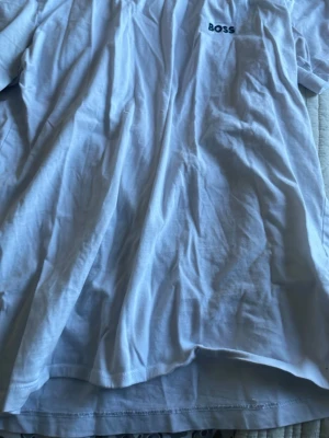 Vit t-shirt från BOSS - Klassisk vit t-shirt från BOSS med diskret logga på bröstet. T-shirten har rund hals och korta ärmar, perfekt för en clean och enkel stil. Tillverkad i mjuk bomull som känns skön mot huden.