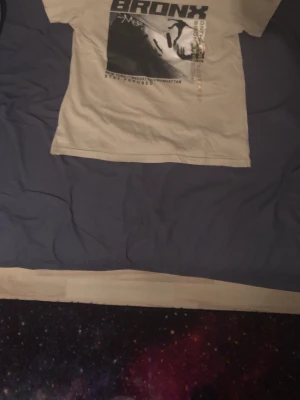 Beige t-shirt med Bronx-tryck Firefly - Snygg beige t-shirt från Firefly med stort svart Bronx-tryck och coolt motiv framtill. Klassisk passform och rund halsringning. Perfekt för dig som gillar streetstyle och vill sticka ut med ett statement-plagg.