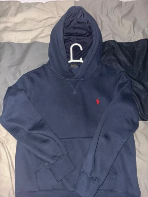 Marinblå hoodie från Polo Ralph Lauren - Snygg marinblå hoodie från Polo Ralph Lauren med klassisk röd logga broderad på bröstet. Hoodien har huva, känguruficka och ribbade muddar vid ärmslut och nederkant. Tillverkad i mjukt bomullsmaterial som är skönt att bära.