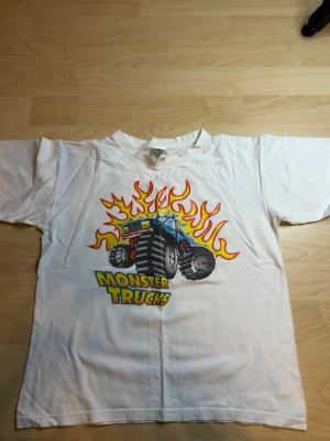 Vit Monster Trucks t-shirt B&C - Säljer en vit t-shirt från B&C Collection med ett färgglatt tryck av en monstertruck och eldflammor på framsidan. T-shirten har korta ärmar och en klassisk passform. Perfekt för dig som gillar coola motiv och streetstyle.