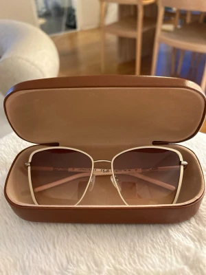 Beige fyrkantiga solglasögon Longchamp - Stilrena solglasögon från Longchamp med fyrkantiga, stora bågar i vit och tunna metallskalmar. Glasen är lätt tonade i brunt och ger en modern och trendig look. Kommer med ett snyggt brunt fodral.