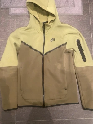Nike hoodie olivgrön med dragkedja - Nike hoodie i två nyanser av olivgrönt med hel dragkedja och huva. Snygg svart Nike-logga på bröstet, fickor framtill och en extra ficka med dragkedja på ärmen. Tillverkad i mjukt sweatshirtmaterial, perfekt för en chill streetwear-stil.
