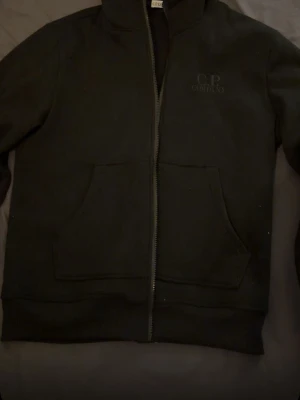 Svart hoodie från C.P. Company - Säljer en svart hoodie från C.P. Company med dragkedja framtill och två stora fickor. Diskret logga broderad på bröstet. Klassisk rak passform och mjukt material som känns skönt mot huden. Perfekt för dig som gillar stilrena och enkla plagg. 