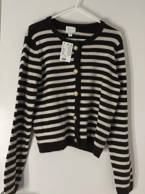 Randig brunvit  kofta från H&M - Snygg mörk brun/svart vit randig kofta från H&M med guldknappar framtill. Koftan har rund halsringning, långa ärmar och ribbade muddar. Perfekt för dig som gillar klassiska mönster med en twist. Materialet är mjukt och stretchigt för extra komfort.