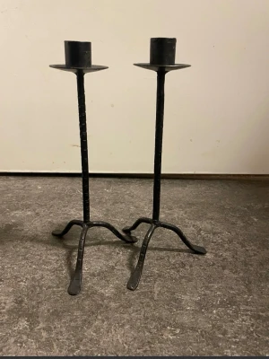 2st Svarta smidesljusstakar ca 25 cm höga  - Två höga ljusstakar i svart smide med industriell känsla. De har tre böjda ben och en enkel, rak design. Perfekta för att skapa en cool och stilren look i rummet. Passar vanliga kronljus.