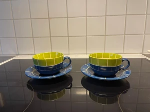 Två retro koppar med fat - Säljer två unika koppar med tillhörande fat i keramik. Kopparna har ett rutigt mönster i limegrönt och blått, med matchande blåa fat och vita linjer. Perfekta för te eller kaffe och ger en färgklick till köket. Säljes som set.