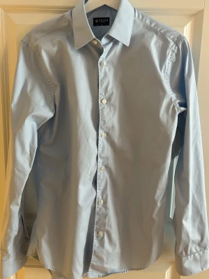 Ljusblå skjorta från Tiger of Sweden - Klassisk ljusblå skjorta från Tiger of Sweden (slim fit) med lång ärm och vita knappar framtill. Skjortan har en stilren krage och är tillverkad i bomull, perfekt för en clean och snygg look.