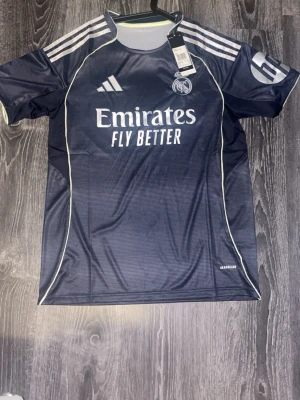 Real Madrid adidas matchtröja mörkblå - Snygg mörkblå Real Madrid matchtröja från adidas med vita detaljer och klubbmärke på bröstet. Tröjan har korta ärmar, rund hals och tre klassiska adidas-ränder på axlarna. Tillverkad i lätt och ventilerande material, perfekt för fotbollsträning eller match.