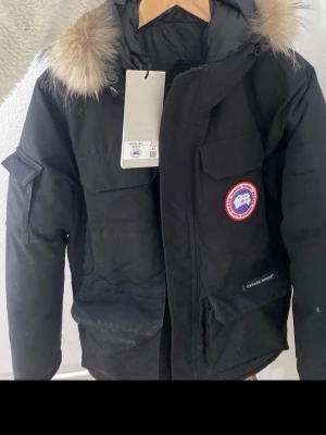 Svart parkas från Canada Goose - Snygg svart parkas från Canada Goose med klassisk patch på bröstet och stor huva med äkta päls. Jackan har flera praktiska fickor och stängs med dragkedja och vindslå. Materialet är polyester och dunfyllning för extra värme. Perfekt för kalla dagar. 100 äkta knappt använd och storlek M