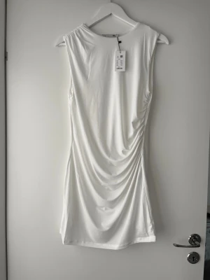 Vit draperad klänning  - Säljer en stilren vit kortklänning från Lager157 med snygg drapering på sidan. Klänningen har ärmlös design och rund halsringning. Tillverkad i ett mjukt och stretchigt material som ger en smickrande passform. Undertyg under klänningen. 