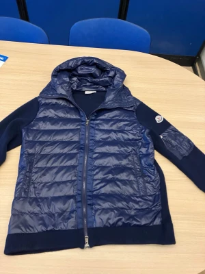 Mörkblå pufferjacka från Moncler - Snygg mörkblå pufferjacka från Moncler med quiltad framsida och huva. Jackan har stickade ärmar och rygg, dragkedja framtill och Moncler-logga på ärmen. Perfekt för kalla dagar och riktigt stilren look.